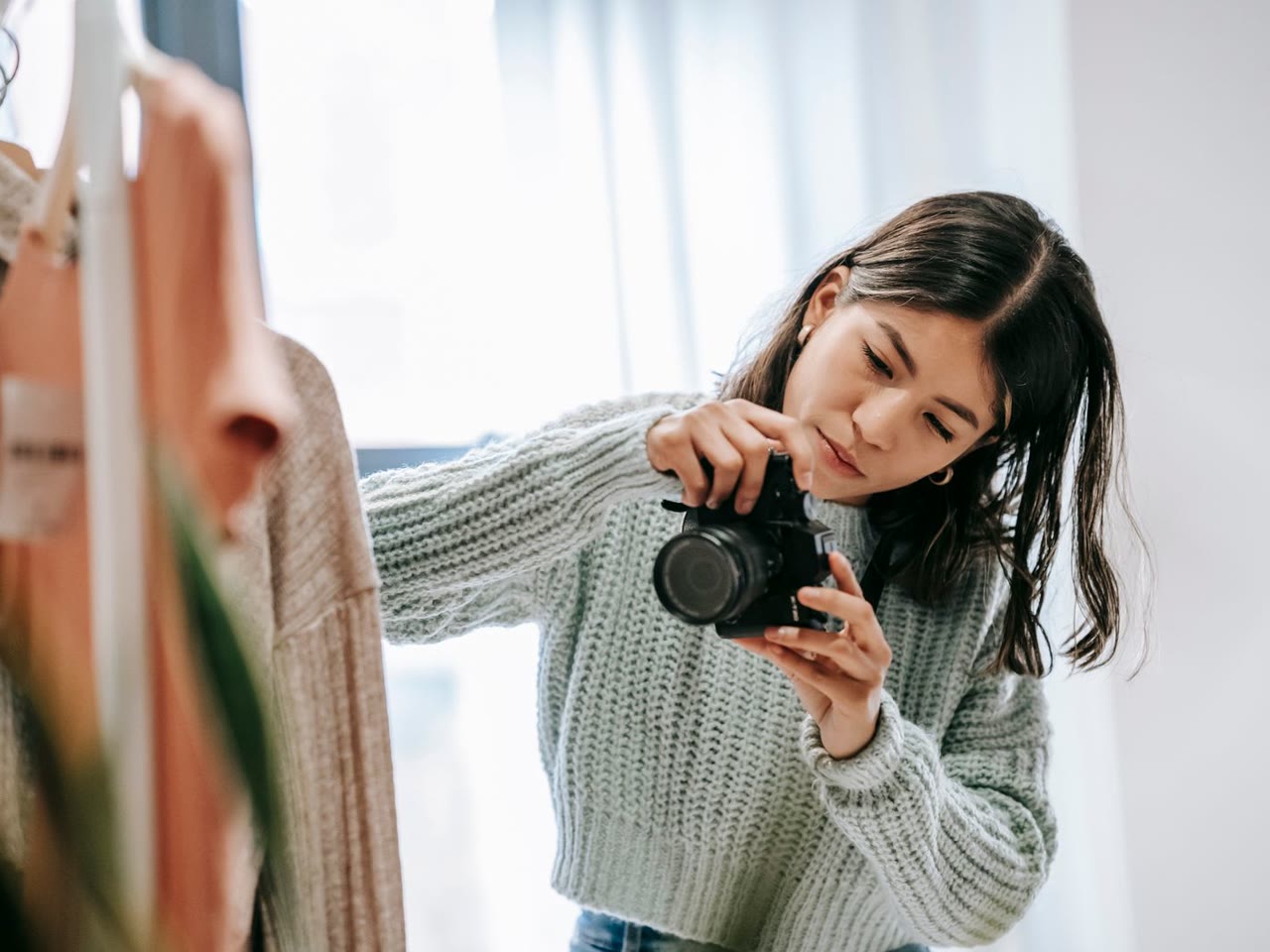 Fotógrafa confere a câmera ao lado de roupas em uma arara, perto de uma janela